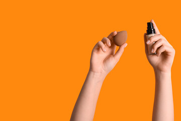 Female hands with makeup sponge and bottle of foundation on orange background