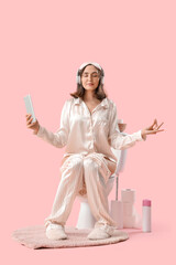 Young woman in pajamas with headphones and tablet computer meditating on toilet bowl against pink...
