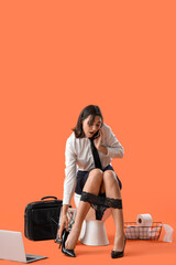 Stressed young businesswoman talking by mobile phone on toilet bowl against orange background