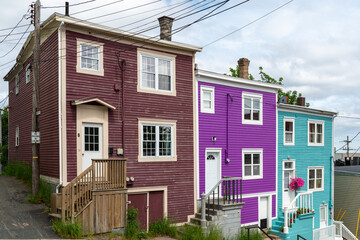 Street view of multiple colorful wooden residential buildings of various colors. The historic houses are row or adjoined. The buildings are on a hill with a narrow sidewalk in front of the old homes.