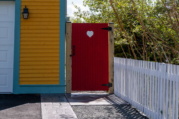 A red wooden gate with a small white painted heart. The garden gate is attached to a white picket fence and a vibrant yellow wooden house with teal colored trim. The yard has tall lush green trees.