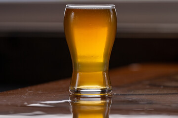 A small curved tulip shaped sample glass of pale ale beer with froth on the top. The liquid alcohol has a vibrant yellow tint. The glass is on a wooden table with a grey background. 