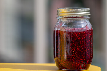 A clear glass jar or crock filled with homemade raspberry breakfast jam. The bottle has a silver...
