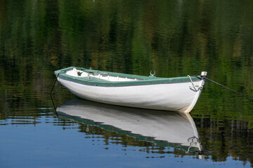 A small white colored wooden traditional dory or fishing vessel with green painted edging. The boat is floating on top of smooth water. The pond reflects the image of the recreational watercraft.