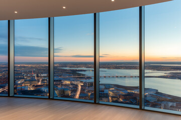View from a high-rise building overlooking a city at sunset. The view is from a room with floor-to-ceiling windows that are framed in black, and the floor is made of light-colored wood