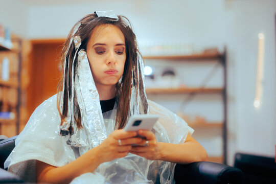 Bored Woman Waiting with Tin Foil in Her Hair Checking her Phone. Impatient girl texting and browsing the internet in a hair salon
