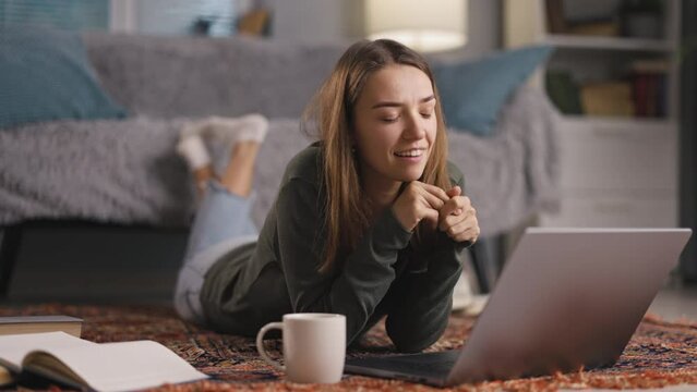 Young positive woman have conversation with laptop remotely, lying on floor with laptop, looking at screen, tells funny stories to friend