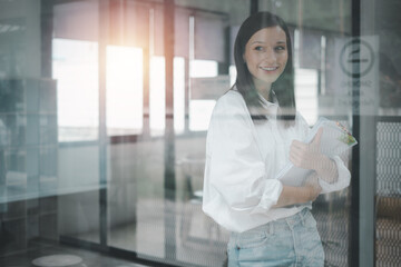 Successful businesswoman standing in creative office and looking out windows. Woman entrepreneur in a coworking space smiling..