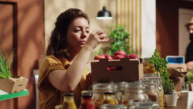 Vegan woman in eco friendly zero waste supermarket counting apples, adding them to shopping basket. Client in local neighborhood grocery shop picking farm grown handpicked fruits