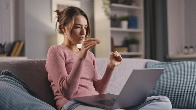 Medium shot of young happy woman video chatting on laptop while staying at home with in evening, sitting on comfortable couch with pillows