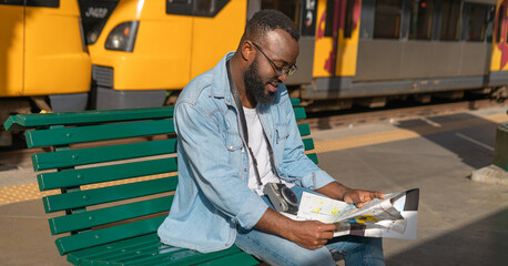 Happy young african american man sitting with map at european terminal train station. Tourist travel by train on vacation time holiday weekend trip.