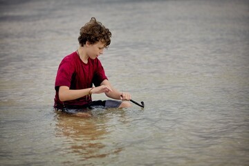 A teenage boy sits in the ocean water and digs with a stick as a play fishing rod