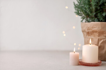 Candles and blurred defocused garland lights, juniper in pot on beige table and empty white wall background. Aesthetic Scandinavian Christmas home decoration, interior design template