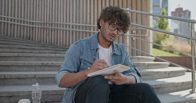 Young indian guy student making notes in a notebook while sitting on a stairs outdoor
