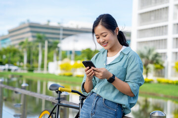 Young Asian woman standing near bicycle. She looks at social media on her cell phone at a park in a...
