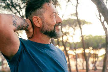 Side view of happy adult man enjoying leisure outdoor activity at the park touching his head and smiling with trees and sunset light in background. People and nature connection. Serene person outside - obrazy, fototapety, plakaty
