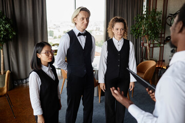 Staff meeting in luxury restaurant with three servers wearing classic uniform and listening to manager in dining room