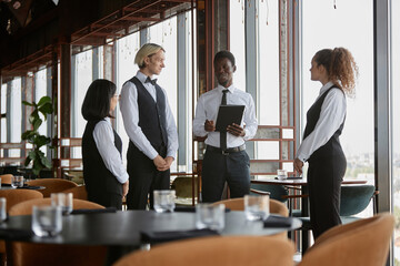 Portrait of smiling Black man as restaurant manager talking to servers wearing classic uniforms during morning meeting