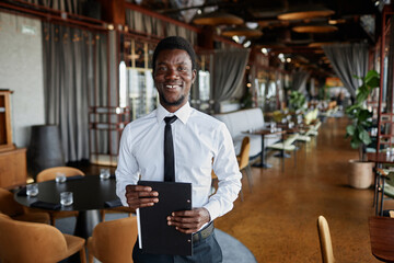 Waist up portrait of Black young man as restaurant manager smiling at camera standing in dining room, copy space - obrazy, fototapety, plakaty