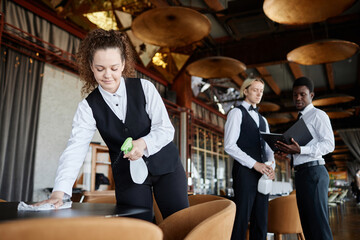 Portrait of smiling young woman as waiter dressed in classic black and white uniform wiping tables...