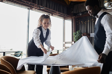 Portrait of curly haired young woman as classic server preparing restaurant for opening and folding tablecloths, copy space