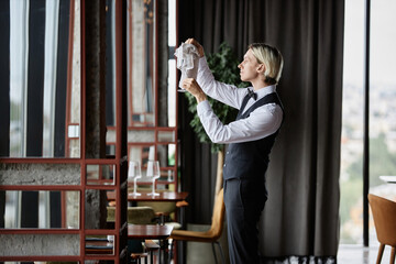 Fototapeta na wymiar Side view portrait of elegant waiter polishing glasses in luxury restaurant interior, copy space