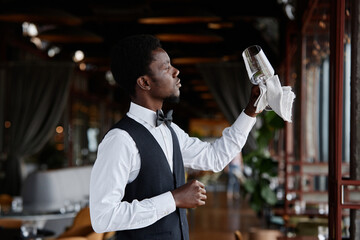 Side view portrait of Black young man as elegant waiter holding crystal glass in restaurant...