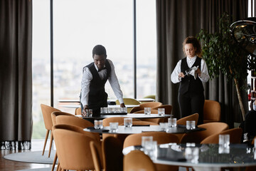 Luxury restaurant staff preparing for grand opening with two elegant servers setting tables in dining room, copy space