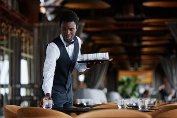 Waist up portrait of elegant Black man as waiter setting tables with crystal glasses preparing for...