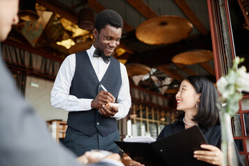 Portrait of Black young man as server taking orders from guests and smiling enjoying work in luxury restaurant, copy space