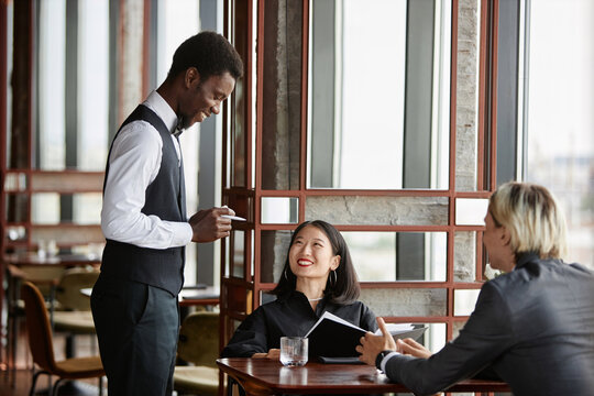Side view portrait of Black young man as server taking orders from couple and smiling in luxury restaurant, copy space