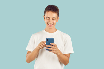 Happy teenager using modern cell phone. Studio portrait adolescent boy in T shirt standing isolated on light blue background holding smartphone, scrolling news feed, looking at funny memes and smiling - obrazy, fototapety, plakaty