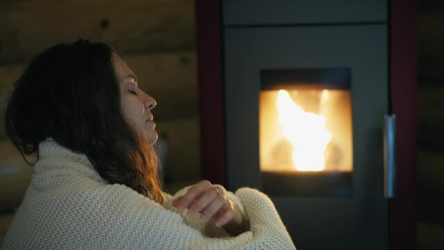 A young Caucasian woman is sitting by an electric pellet fireplace wrapped in a blanket.