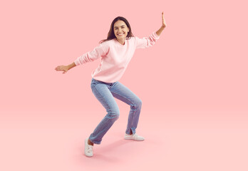 Cheerful funny young woman having fun fooling around dancing on pink background in studio. Caucasian brunette woman in casual sweatshirt and jeans. People lifestyle concept. Full length. Isolated.