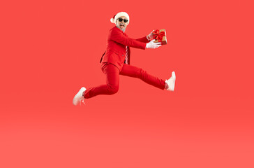 Full length portrait of crazy man wearing Santa style holding Xmas gift present jumpinh in air on red background. He is smiling and looking at camera. Christmas winter holiday advertisement banner.