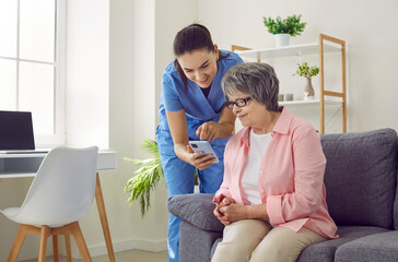 Elderly care. Friendly female caregiver shows elderly woman how to use modern mobile applications to monitor her health. Nurse and senior woman in nursing home are looking at mobile phone screen. - obrazy, fototapety, plakaty