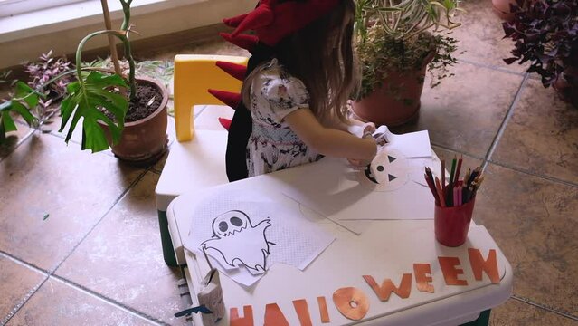 Little girl making Halloween home decorations sitting at wooden table, painting pumpkins and making paper cuttings