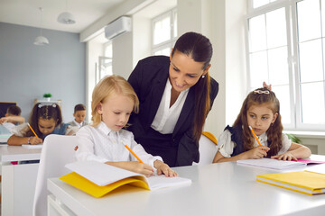 Friendly female teacher observes and helps little boy with his schoolwork during lesson. Smiling woman leans over and looks at schoolboy writing in his notebook. School concept.