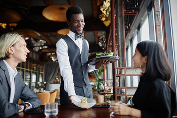 Portrait of smiling waiter bringing food on tray to guests enjoying dinner in luxury restaurant