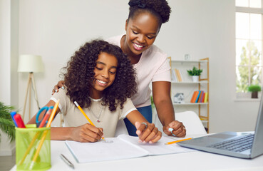 Happy, cheerful family doing school homework. Supportive parent helping child with homework. Smiling African American mother and daughter writing and drawing in notebook at desk with laptop computer - obrazy, fototapety, plakaty