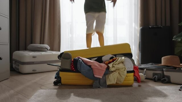 Boy Happily Jumps On Packed Suitcase. Legs Feets Of Small Child, Messy Chaotically Packed Suitcase With Clothes Sticking Out. Kid On Packed Suitcase Experiences Storm Of Emotions Anticipating Vacation