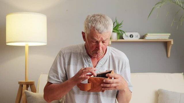 Elderly mature gentleman sitting on couch with unfilled wallet devoid of funds for expenditure conveying sorrow and disillusionment expressing sadness.