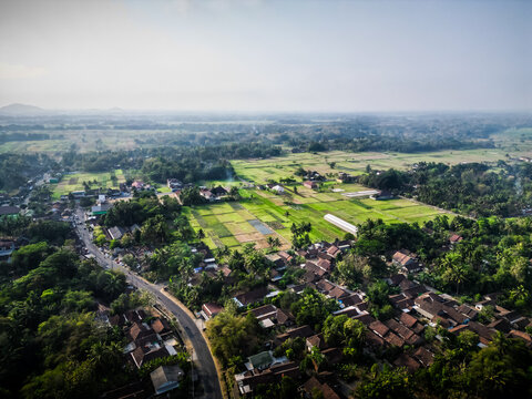 Aerial view of human settlement buildings expansion near paddy rice fields in the morning sunlight. Kulon Progo rural village landscape. Population density is displacing agricultural land and forests.