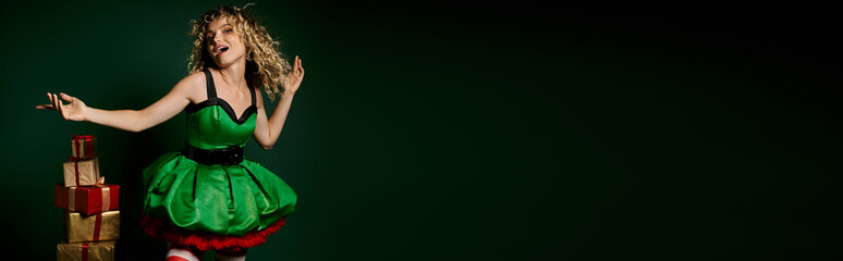 Naklejka premium jolly woman in elf costume gesturing with arms posing near pile of gifts on green backdrop, banner