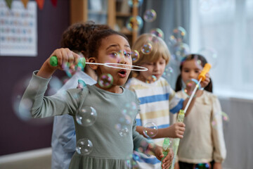 Group of joyful little kids playing with soap bubbles in preschool and laughing happily