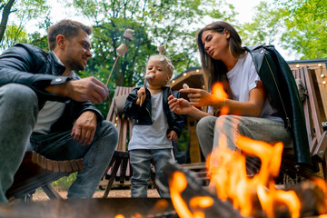 Cute young family dad mom and daughter are sitting together by fire in the forest and roasting...