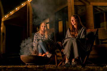 Obraz premium Girlfriends in blankets sit in the evening by the fire near country wooden house in the forest