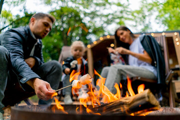 Cute young family dad mom and daughter are sitting together by fire in the forest and roasting...