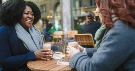 Young multiracial women having coffee break at vintage bar outdoor during winter time - Cozy beverage and lifestyle concept - Focus on african girl face