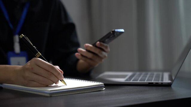 Female businessman use a smartphone and take notes in the notebook at the office desk in the morning.